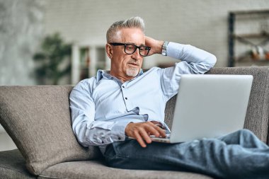 Çekici, olgun bir adam evinde rahat bir kanepede otururken dizüstü bilgisayarını karıştırıyor. Ev ve yaşam tarzı kavramı