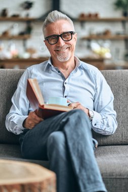 Oturma odasındaki rahat kanepede dinlenen neşeli, gri saçlı yetişkin bir adam elinde bu ve bir fincan kahve tutarken. Yurtiçi yaşam tarzı kavramı