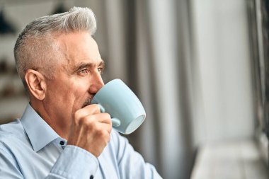 Evinin penceresinin dışındaki şehrin manzarasına bakarken beyaz saçlı yetişkin bir adamın kahve içişini yakından çek. Boş zaman ve yaşam tarzı konsepti