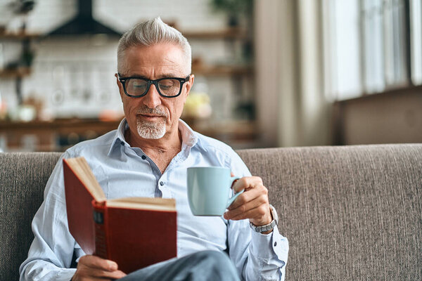 Waist up of mature man reading book while sitting on sofa in his living room and holding cup of coffee. Domestic lifestyle concept. Copy space