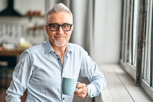 Evinde kahve tutarken pencerenin kenarına yaslanan mavi gömlekli yakışıklı, olgun bir adamın belini kaldır. Boş zaman ve yaşam tarzı konsepti