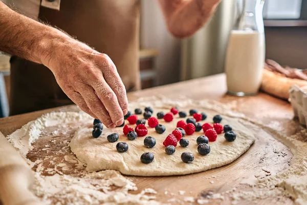 Cropped photo of chef cooking a pie with berries while putting raspberries and blueberries on the dough. Baking process concept