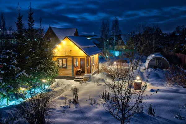 Illuminated at night with spotlights this wooden guest-house  banya framed by spruce and birch trees after a heavy snowfall in winter twilight. Moscow Region, Russia.