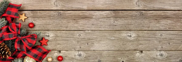 Christmas corner border of ornaments, branches and buffalo plaid check ribbon. Top down view on a rustic wood banner background.