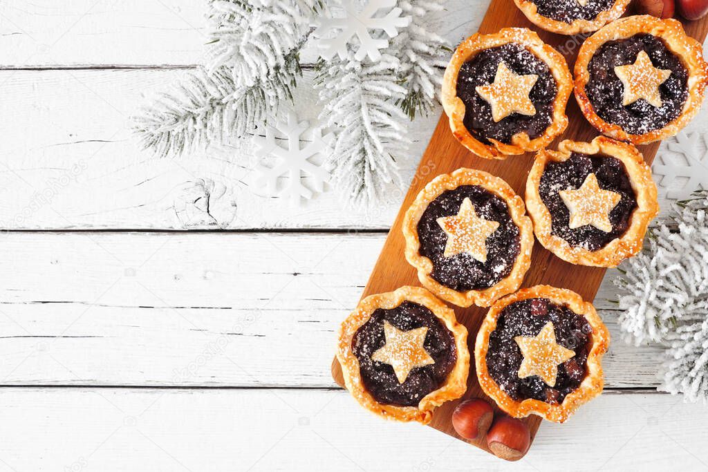 Christmas mincemeat tarts on a serving board. Above view corner border over a white wood background. Copy space.