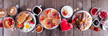Sevgililer Günü ya da Anneler Günü brunch masası sahnesi. Koyu renk ahşap afiş arkaplanda yukarıdan görünüyor. Kalp şeklinde krepler, yumurtalar ve çeşitli aşk temalı yemekler..