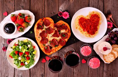 Ev yapımı Sevgililer Günü yemeği. Koyu ahşap arka planda, görüş masasının üstünde. Kalp şeklinde pizza, makarna, şarap, peynir tabağı ve tatlılar..