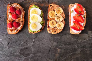 Çeşitli protein ve meyvelerle hazırlanmış sağlıklı tost. Fotokopi alanı olan koyu bir arkaplanda en üstteki görünüm. Krem peynirli ahududu, avokado yumurtası, fıstık ezmeli muz chia, çilekli yoğurt..
