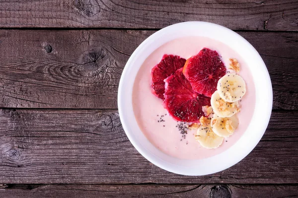 Healthy blood orange and banana smoothie bowl with walnuts and chia seeds. Overhead view on a dark wood background.