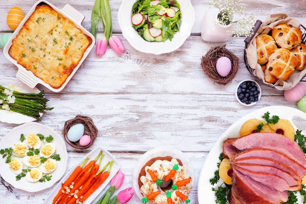 Classic Easter ham dinner. Above view frame on a white wood background with copy space. Ham, scalloped potatoes, vegetables, eggs, hot cross buns and carrot cake.