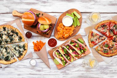 Sağlıklı bitki tabanlı fast food masası sahnesi. Beyaz ahşap arka planda üst manzara. Karnabahar hamurlu pizzalar, fasulye burgerler, mantarlı tacolar, dolmalık nacholar ve karnabahar kanatları ve tatlı patates kızartmaları..