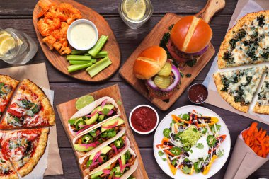 Sağlıklı bitki tabanlı fast food masası sahnesi. Ahşap arka planda genel manzara. Karnabahar hamurlu pizzalar, fasulye burgerler, mantarlı tacolar, dolmalık nacholar ve karnabahar kanatları ve tatlı patates kızartmaları..