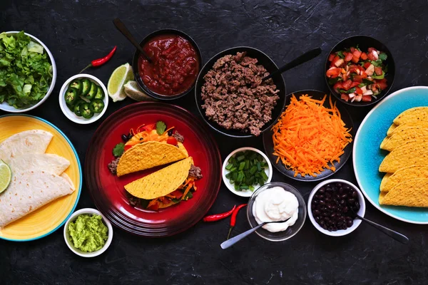 Taco bar table scene with a variety of ingredients. Overhead view on a dark slate background. Mexican food buffet.