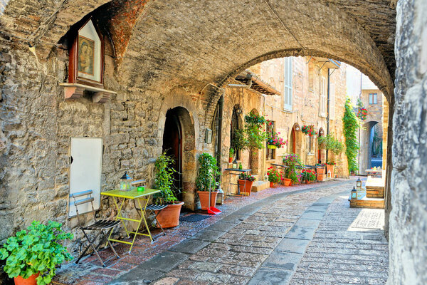 Beautiful arched street in the medieval old town of Assisi with flowers and restaurant tables, Italy
