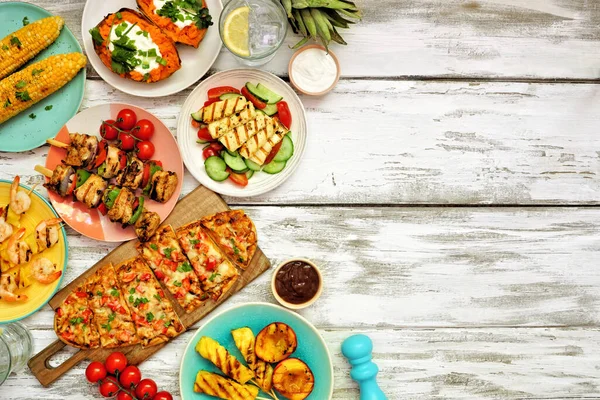 Summer BBQ grill side border over a white wood background. Chicken and shrimp skewers, flatbread, stuffed sweet potato, grilled fruit, corn and salad. Overhead view with copy space.