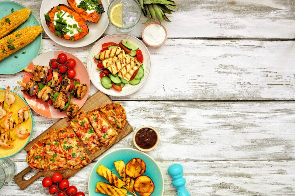 Summer BBQ grill side border over a white wood background. Chicken and shrimp skewers, flatbread, stuffed sweet potato, grilled fruit, corn and salad. Overhead view with copy space.