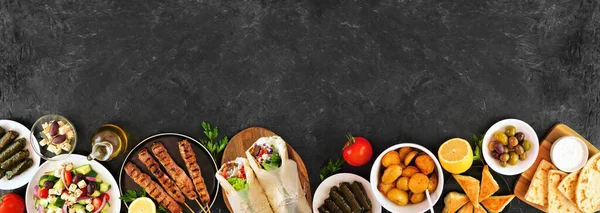 Greek food bottom border, top view on a dark banner background. Souvlaki, gyros wraps, salad, spanakopita, dolmades, pita and lemon potatoes. Copy space.