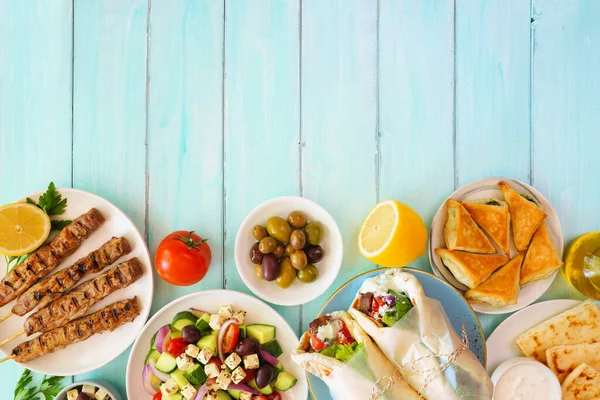 Greek food bottom border, top view on a blue wood background. Gyros wraps, souvlaki, salad, spanakopita, pita and tzatziki. Copy space.