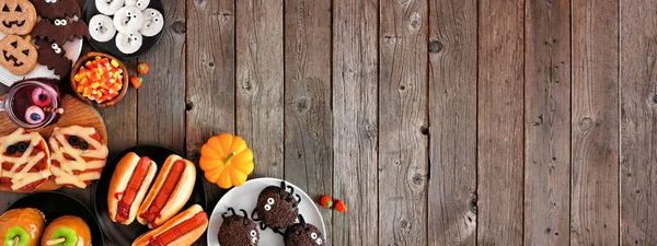 Halloween party food corner border over a rustic wood background with copy space. Top down view. Spooky mummy pizzas, finger hot dogs, caramel apples, cupcakes, candy, cookies and donuts.