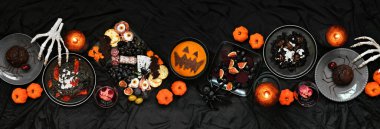 Cadılar Bayramı yemek masası sahnesi koyu siyah bir afişin üzerinde. Manzaranın üstünde. Ürkütücü şarküteri tahtası, siyah risotto ve makarna, örümcek pastası, balkabağı çorbası ve elma kafatası şarabı.