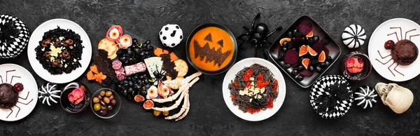 Cadılar Bayramı yemek masası sahnesi siyah afiş arkaplanının üstünde. Manzaranın üstünde. Ürkütücü şarküteri tahtası, siyah risotto ve makarna, örümcek pastası, balkabağı çorbası, salata ve elma kafatası şarabı..