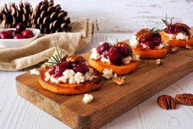 Kızarmış sonbahar tatlı patates crostini aperatifleri peynir, kızılcık ve fındıklı ahşap tabakta. Beyaz ahşap arka planlı masa sahnesi.