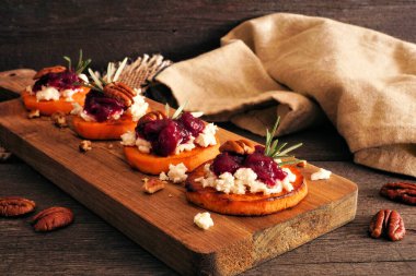Sonbahar tatlı patates crostini aperatifleri peynirli, kızılcıklı ve cevizli ahşap tabakta. Koyu ahşap arka planlı masa sahnesi.