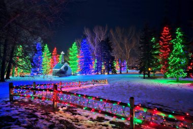 Noel ışıkları sahnesi, ahşap çitlerle kaplı, karla kaplı bir ormanda renkli ışıklarla parlayan Noel ağaçları arasında bir kiliseye açılıyor.