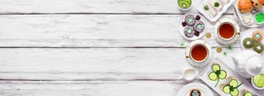 St Patrick's Day tea party side border against a white wood banner background. Assortment of green shamrock theme finger sandwiches, fruits, scones and sweet desserts. Top down view. Copy space.