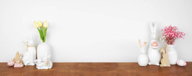 Ahşap bir rafta paskalya dekoru. Paskalya tavşanları, çiçekler, duvardaki beyaz pankartta duran yumurtalar. Boşluğu kopyala.