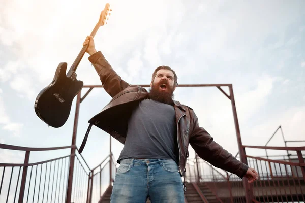 bearded stern male guitarist with gray hair in a brown leather jacket and blue jeans holds an electric guitar in his hand and shouts against the background of the pedestrian crossing stairs