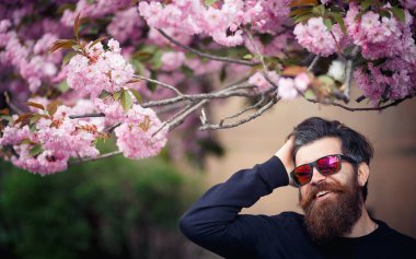 Uzun bıyıklı, gri saçlı, parlak gözlüklü, koyu renk kazaklı ve baharda kiraz çiçeklerinin yanında beyzbol şapkası takan ciddi bir sakallı adam. Dikey fotoğraf