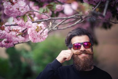 Koyu renk kazaklı, gri saçlı, parlak gözlüklü ve beyzbol şapkalı neşeli sakallı bir adam baharda kiraz çiçeklerinin yanında bıyığını tutuyor ve gülüyor..