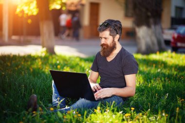 Uzun siyah saçlı, tişörtlü ve kot pantolonlu sakallı bir zampara paradaki yeşil çimlere dizüstü bilgisayarla oturmuş öğle yemeği sırasında internetten kızlarla iletişim kuruyor.