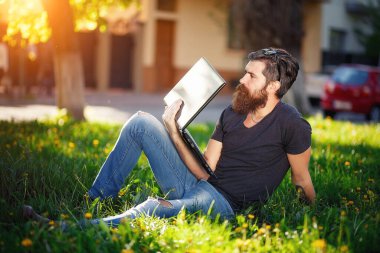 İş adamı, uzun sakallı, gömlekli, koyu gri saçlı ve kot pantolonlu bir iş adamı, parktaki yeşil çimlerde dizüstü bilgisayarla oturuyor ve kâra bakıyor.