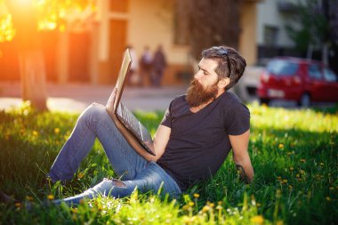 Uzun sakallı, gömlekli ve kot pantolonlu koyu gri saçlı sakallı bir erkek programcı parktaki yeşil çimlerde dizüstü bilgisayarla oturuyor. İnternet 'te serbest çalışma