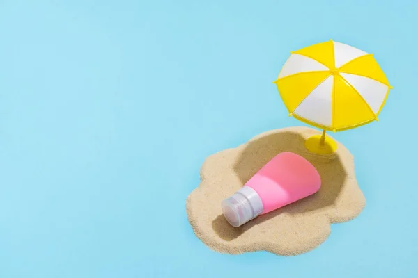 Summer concept. Sunscreen cream on sand and sun umbrella on blue background. Skin care concept in summer.