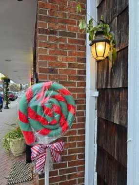 Açık hava dekorasyonunda kırmızı ve yeşil bükülmüş plastiğe sarılı ve çizgili bir yayla bağlanmış büyük bir lolipop kullanılıyor. Beyaz bir çubuğa bağlanmış ve yanan bir fenerin altında, tuğla bir duvara karşı sergilenmiş.