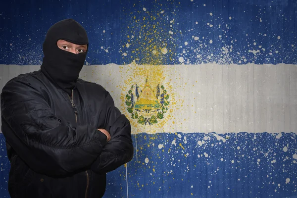 Hombre peligroso en una máscara de pie cerca de una pared con la bandera nacional pintada del salvador — Foto de Stock