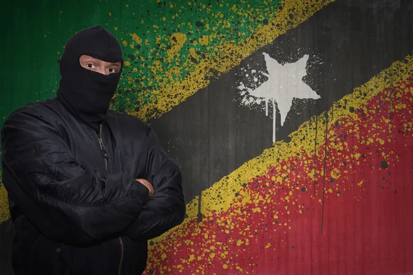 Uomo pericoloso in maschera in piedi vicino a un muro con bandiera nazionale dipinta di gattini santi e nevis — Foto Stock