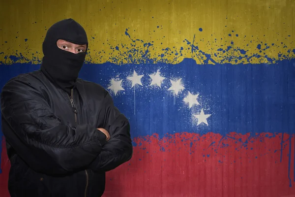 Hombre peligroso en una máscara de pie cerca de una pared con la bandera nacional pintada de venezuela — Foto de Stock
