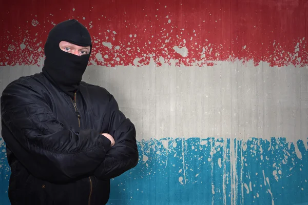Hombre peligroso en una máscara de pie cerca de una pared con la bandera nacional pintada de luxembourg — Foto de Stock