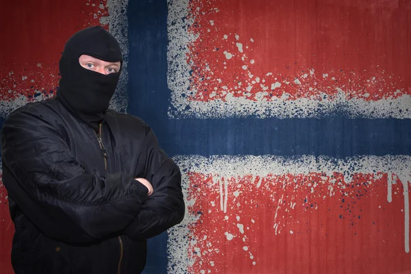 Niebezpieczny człowiek w masce stojącej w pobliżu ściany z namalowaną narodową flagą Norwegii — Zdjęcie stockowe
