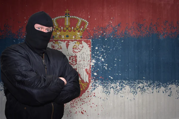 Niebezpieczny człowiek w masce stojącej w pobliżu ściany z namalowaną narodową flagą Serbii — Zdjęcie stockowe