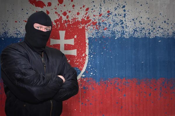 Niebezpieczny człowiek w masce stojącej w pobliżu ściany z namalowaną narodową flagą Słowacji — Zdjęcie stockowe