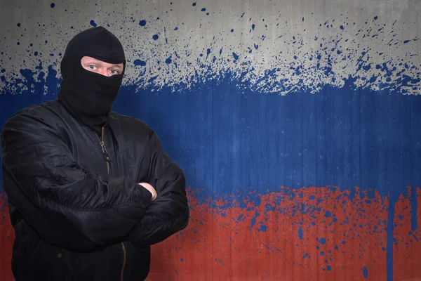 Опасный человек в маске, стоящий у стены с раскрашенным национальным флагом России — стоковое фото