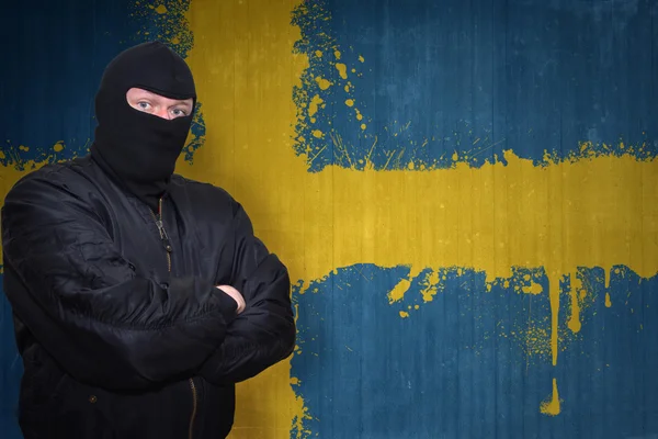 Niebezpieczny człowiek w masce stojącej w pobliżu ściany z namalowaną narodową flagą Szwecji — Zdjęcie stockowe