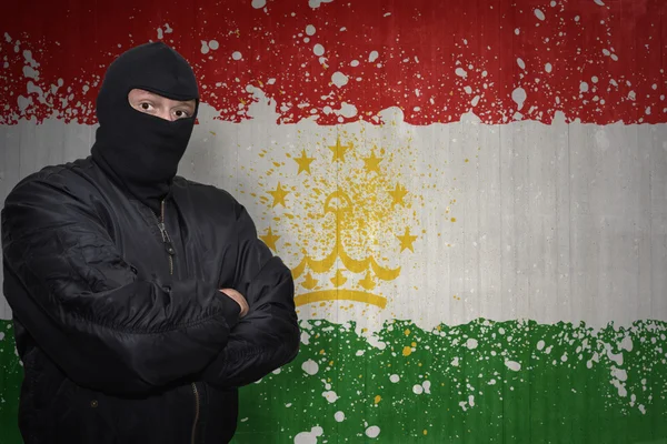 Om periculos într-o mască stând lângă un zid cu steagul național pictat al tajikistanului — Fotografie, imagine de stoc