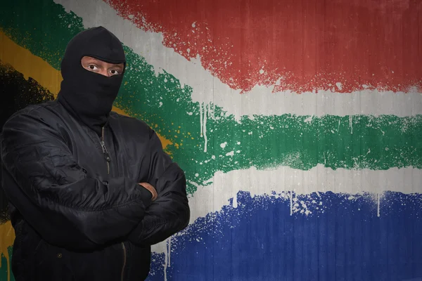 Hombre peligroso en una máscara de pie cerca de una pared con la bandera nacional pintada de Sudáfrica —  Fotos de Stock