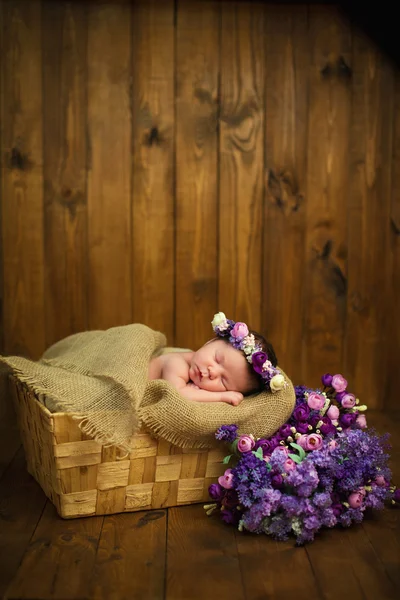 Newborn baby girl with a wreath in a wicker basket with a bouquet of purple wild flowers — Stock Photo, Image